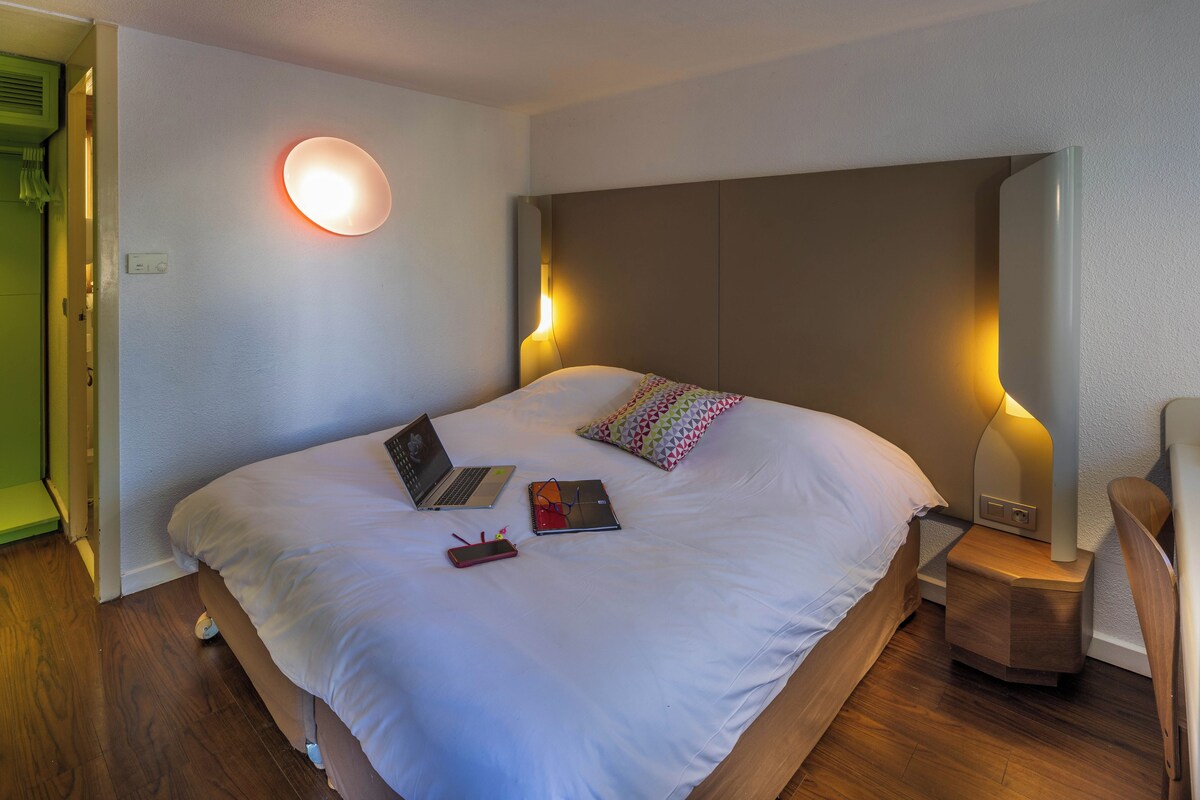 A neatly made bed with a laptop, a smartphone, and a book on it. There is a bedside table with a lamp and a chair.