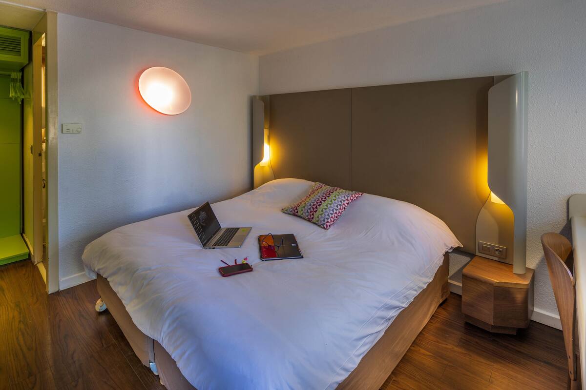 A neatly made bed with a laptop, a smartphone, and a book on it. There is a bedside table with a lamp and a chair.