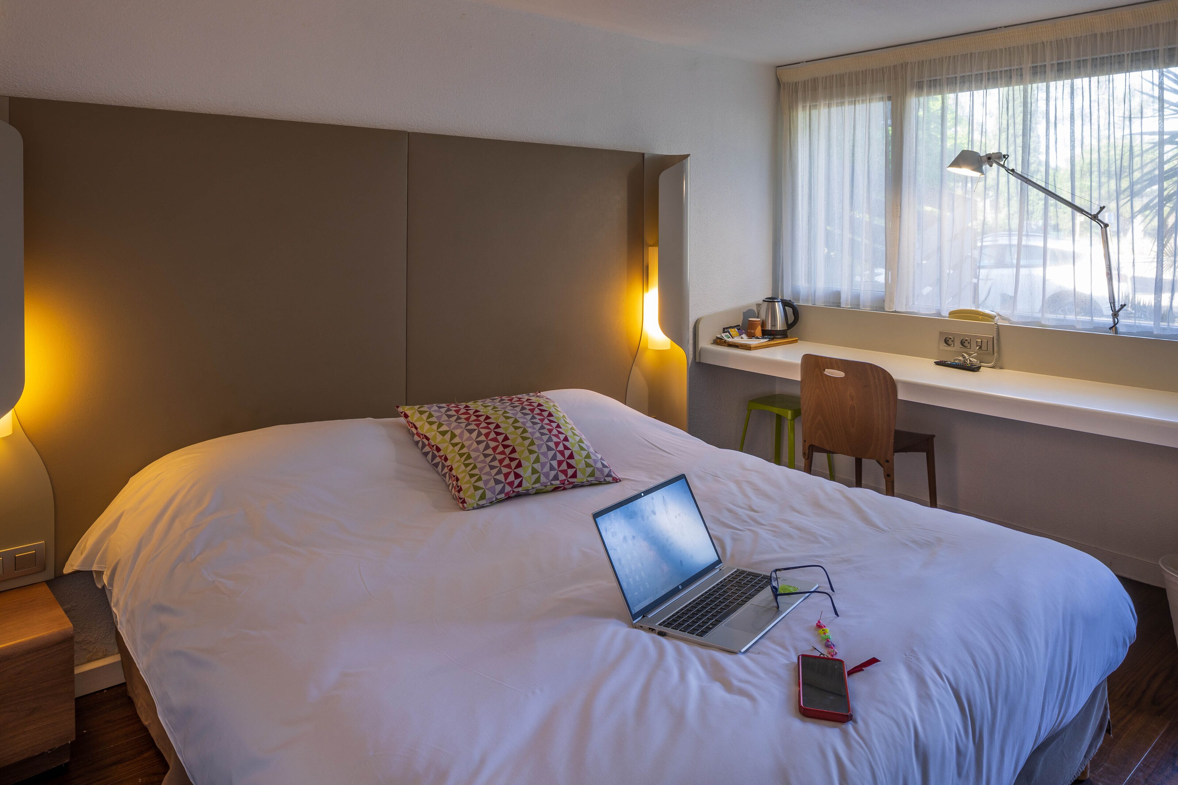 A hotel room with a bed, a laptop on the bed, a desk with a chair, and a window with curtains.