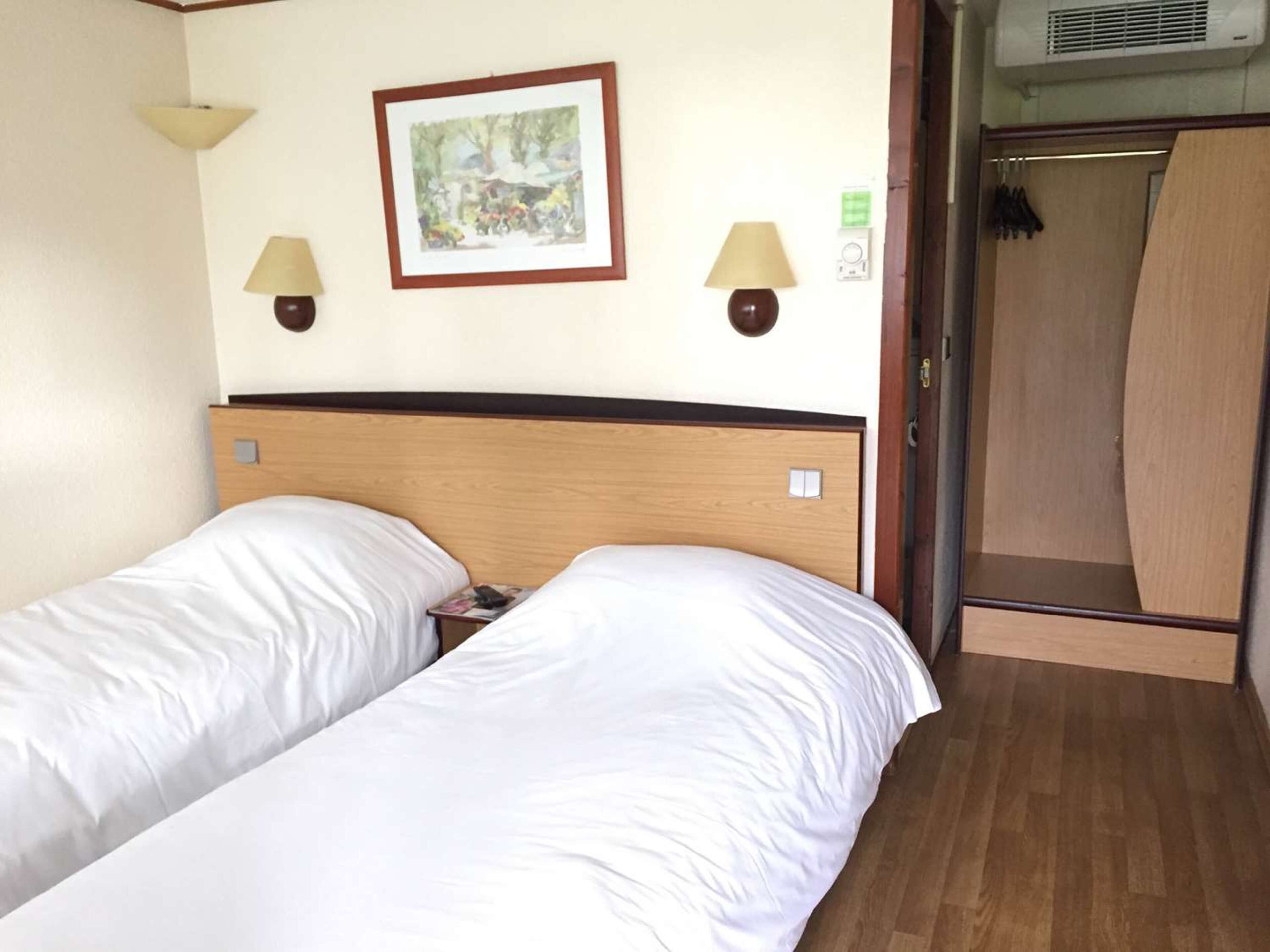 A hotel room with two single beds, a wooden headboard, two wall-mounted lamps, and a framed picture on the wall.
