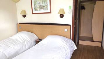 A hotel room with two single beds, a wooden headboard, two wall-mounted lamps, and a framed picture on the wall.