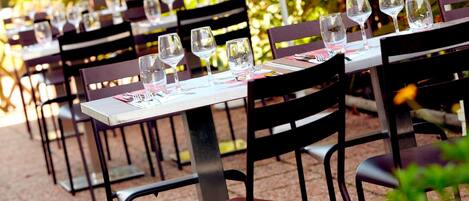 Mesas de comedor al aire libre con sillas, dispuestas con copas de vino y cubiertos, rodeadas de follaje.