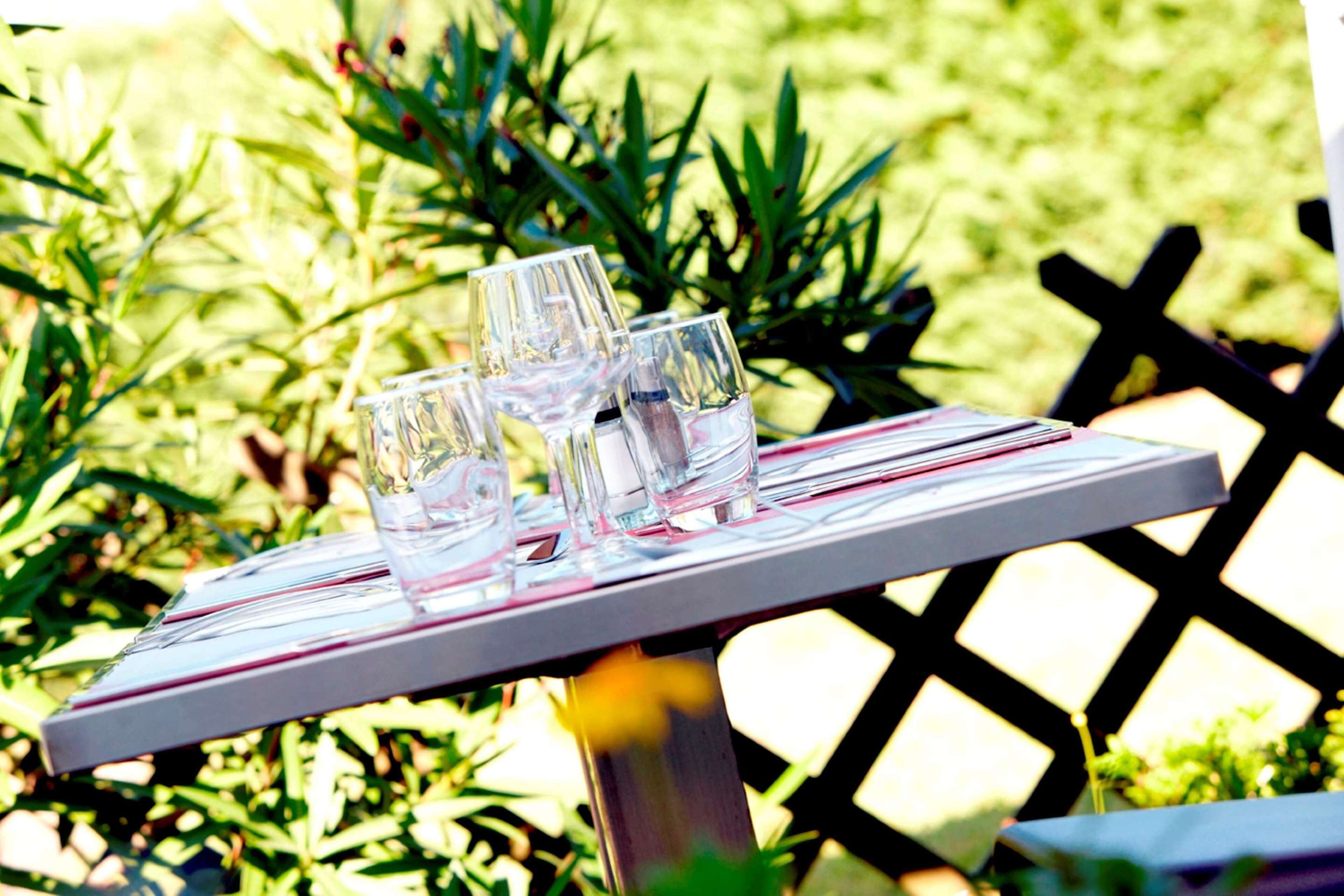 Une table avec quatre verres transparents et un menu, installée à l’extérieur, avec de la verdure en arrière-plan.
