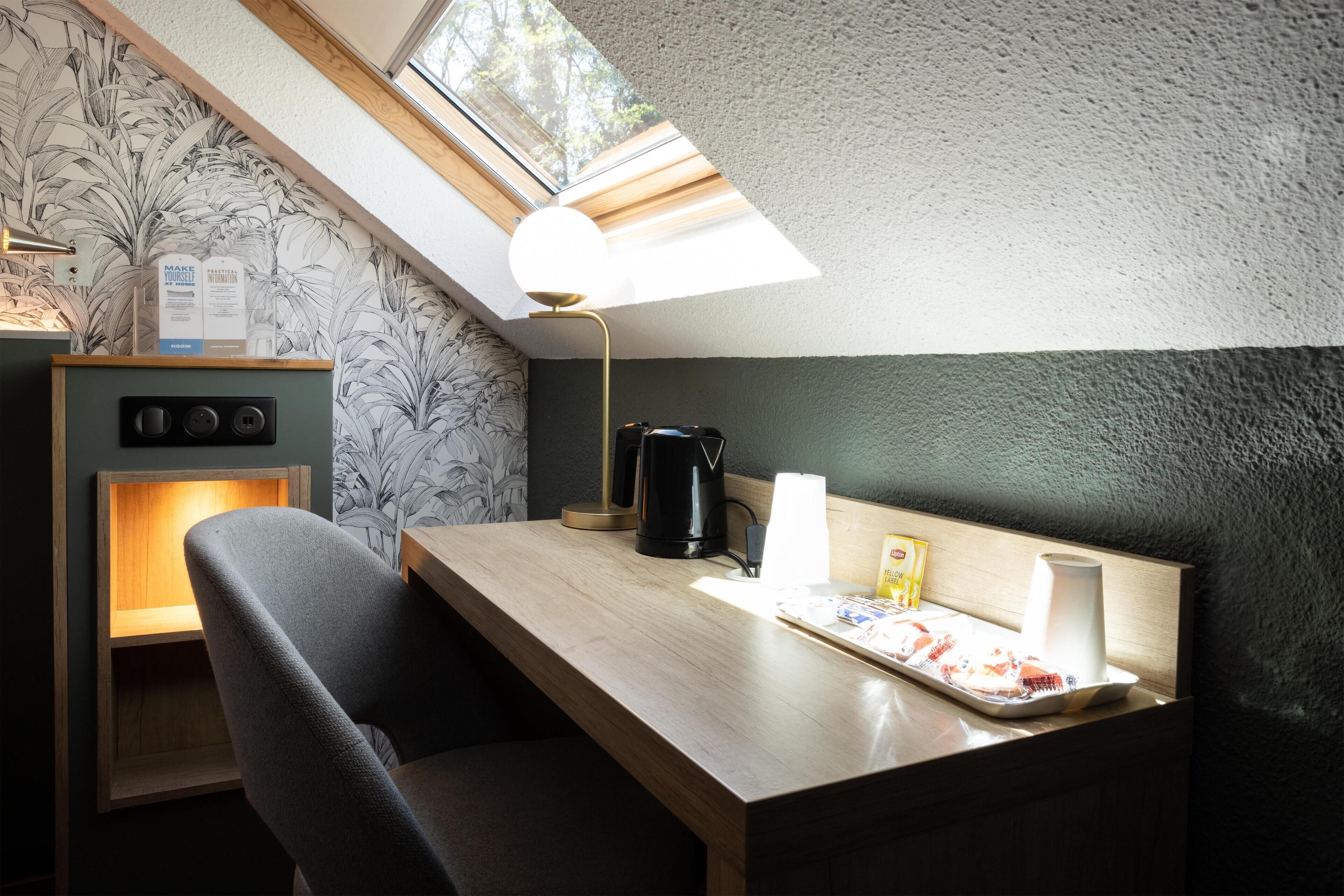 Une petite pièce avec un puits de lumière, un bureau avec une lampe, une chaise, une machine à café et un plateau avec de la nourriture et une tasse.
