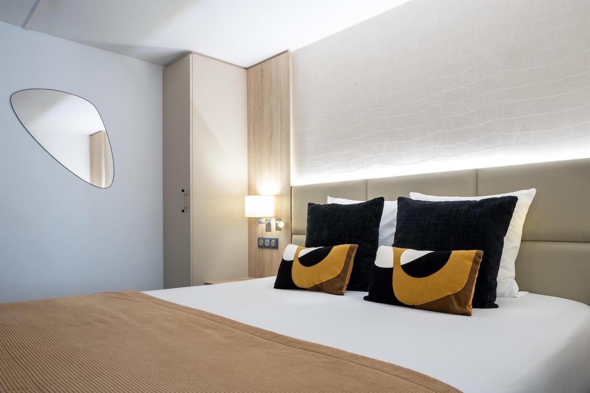 Une chambre d’hôtel comprenant un lit, une table de chevet, une commode, un miroir fixé au mur et une fenêtre avec un luminaire.