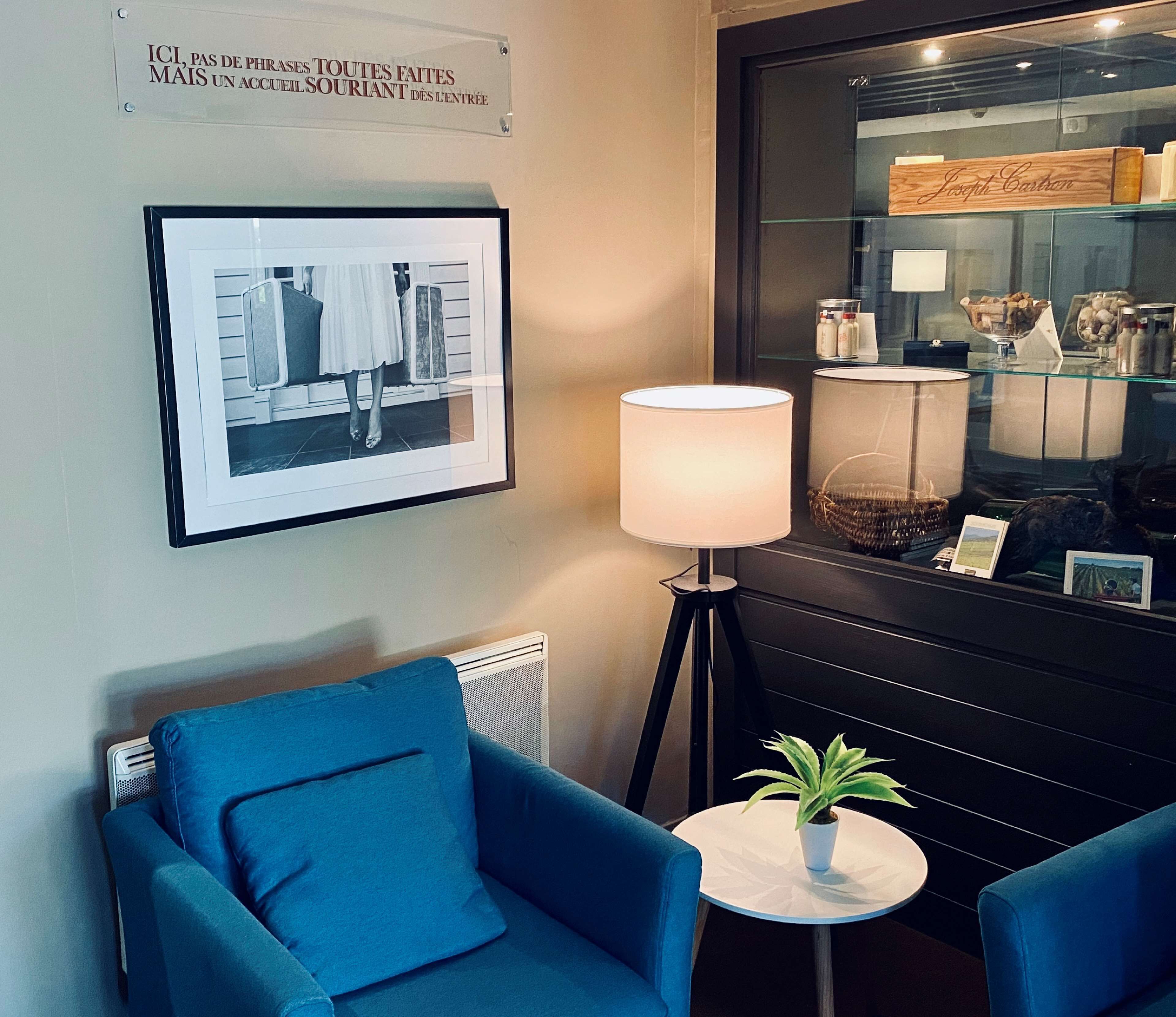 A hotel room with a framed photograph on the wall, a standing lamp, a small round table with a potted plant, and two blue armchairs.