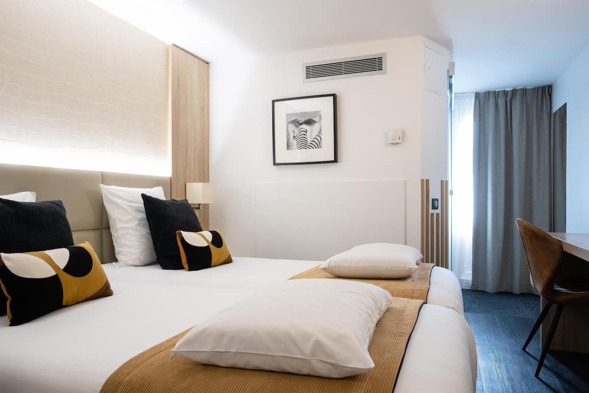 Une chambre d’hôtel avec un lit, un bureau, une chaise, un tableau au mur et une fenêtre avec des rideaux.