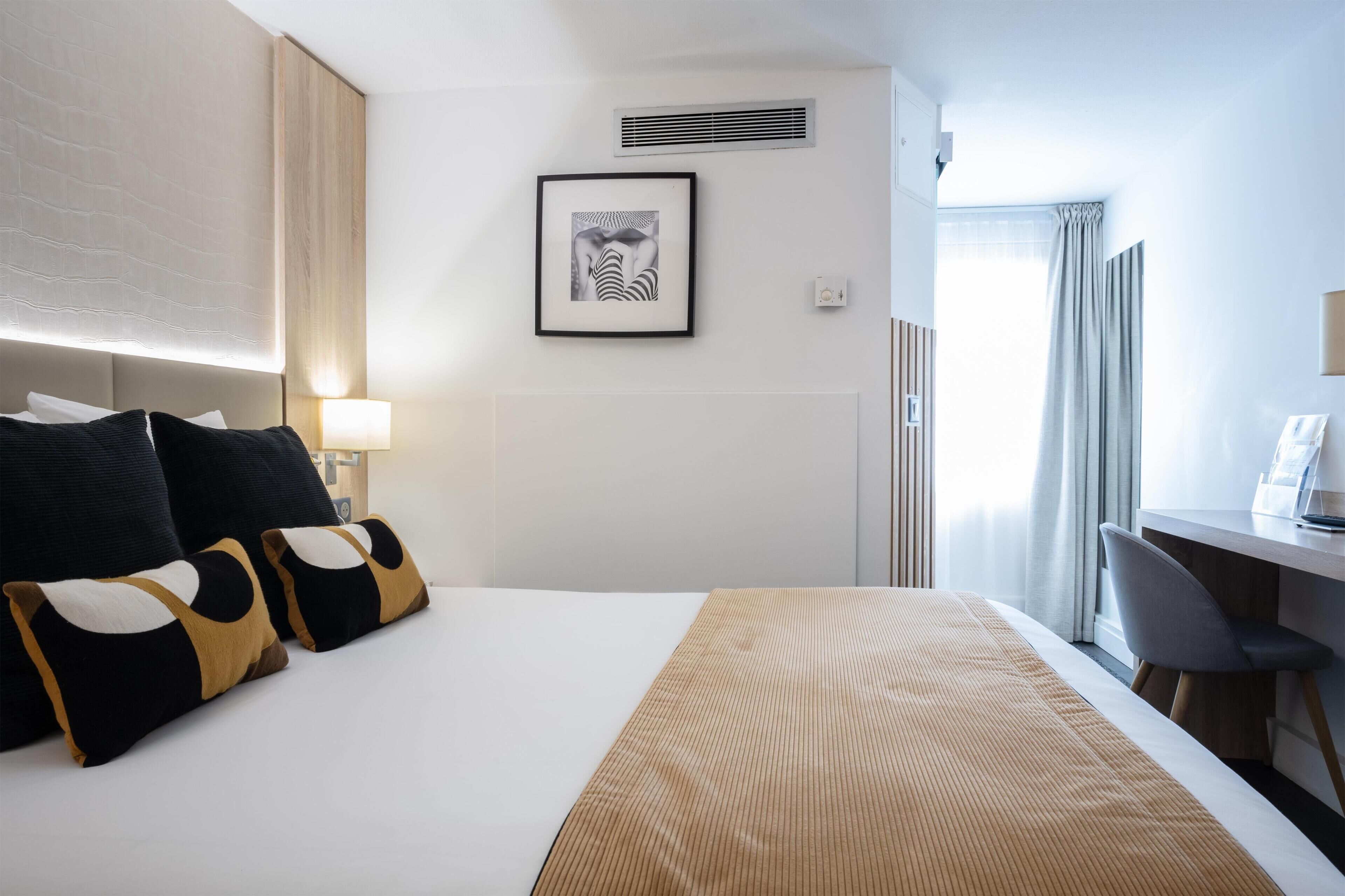 A hotel room with a bed, a desk, a chair, a framed picture on the wall, and a window with curtains.