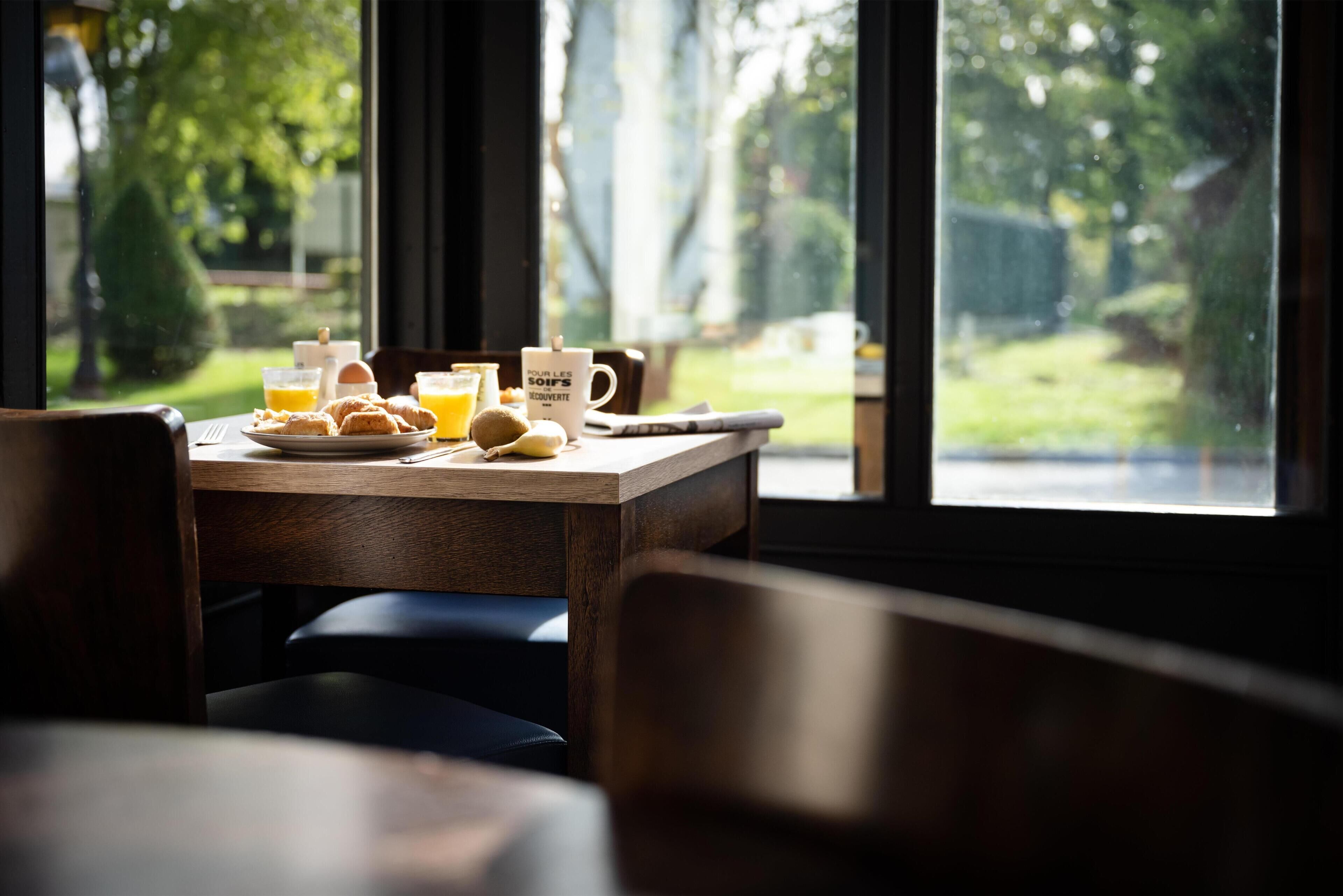 Una mesa de desayuno con jugo de naranja, una taza y pasteles, con vistas a un jardín verde a través de amplias ventanas.