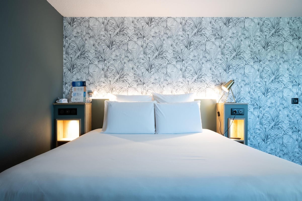 Un lit bien fait, avec du linge de lit blanc, deux tables de chevet avec des lampes et un motif floral sur le papier peint du mur.