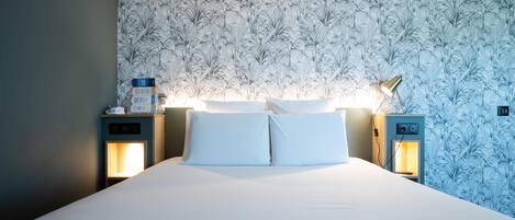 Un lit bien fait, avec du linge de lit blanc, deux tables de chevet avec des lampes et un motif floral sur le papier peint du mur.