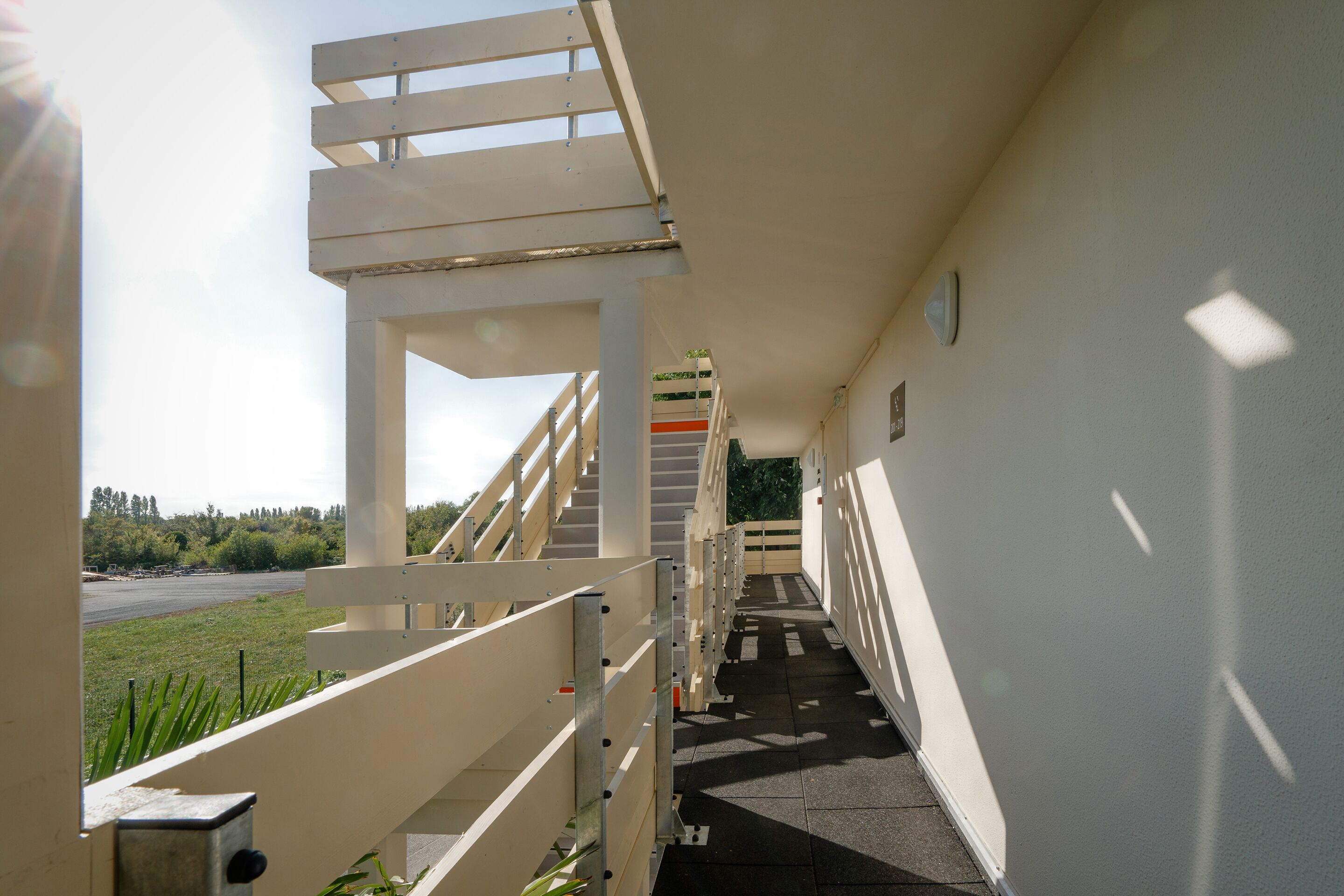 A corridor with a staircase leading to an upper level, a view of a road and greenery outside, and a building with a white exterior.
