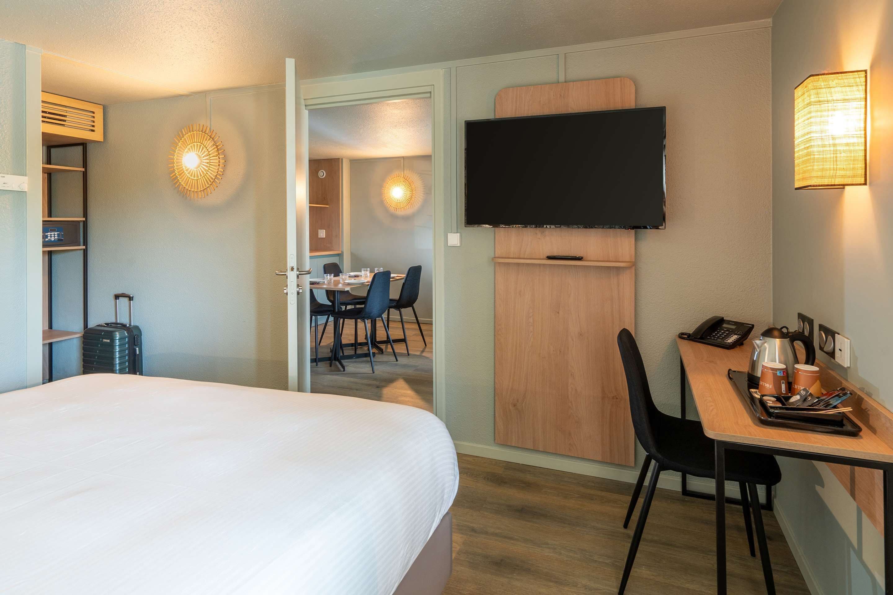 A hotel room with a bed, a desk with a chair, a television, and a dining area in the background.