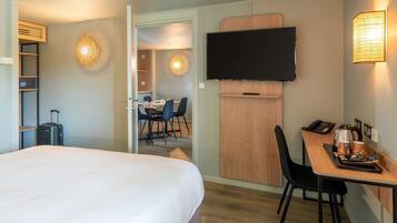 A hotel room with a bed, a desk with a chair, a television, and a dining area in the background.