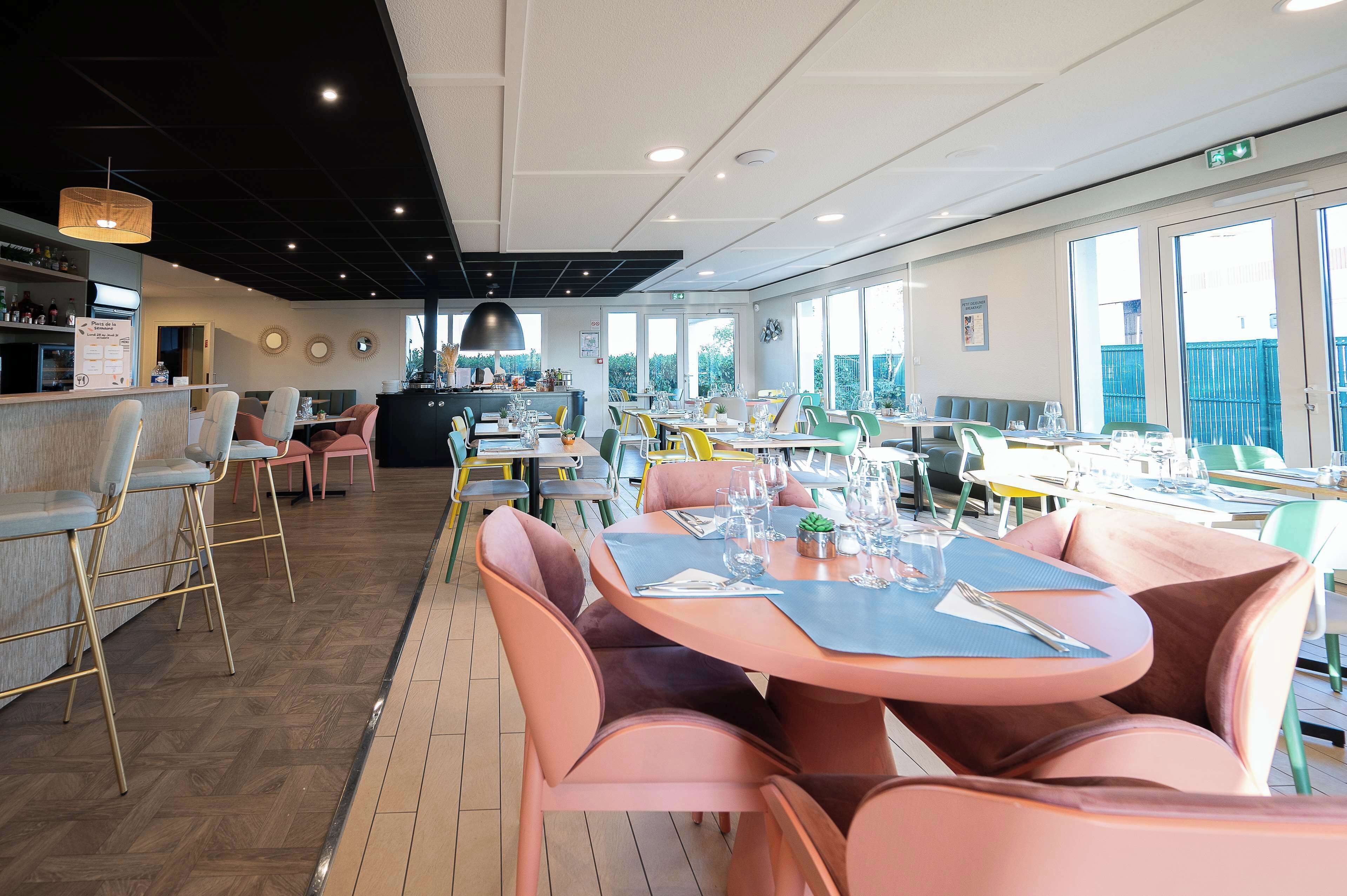 A modern restaurant with round tables and cushioned chairs, a bar area, and large windows offering a view of the outdoors.