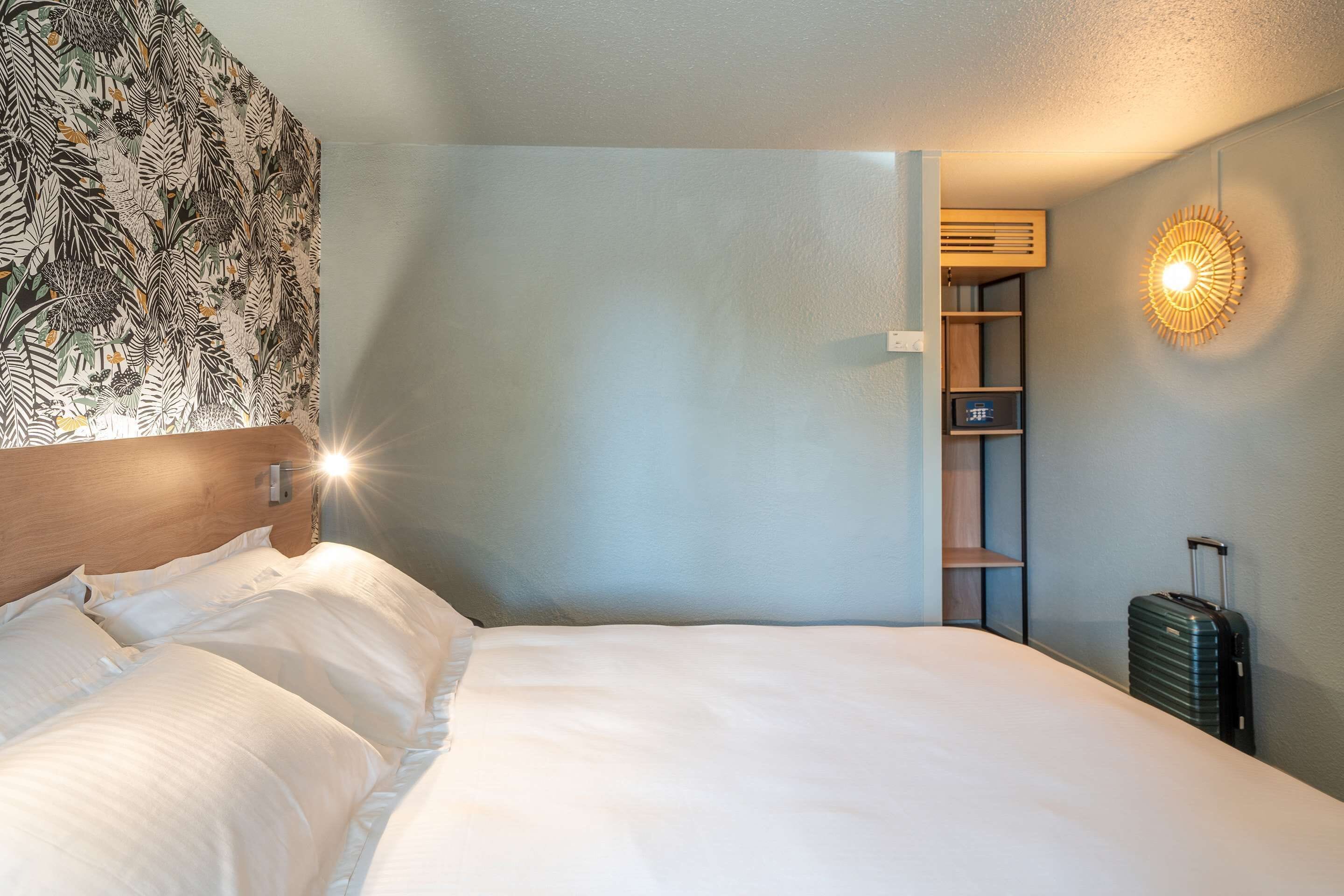Habitación de hotel con cama, cabecera con diseño de hojas, lámpara de pared, armario y maleta.