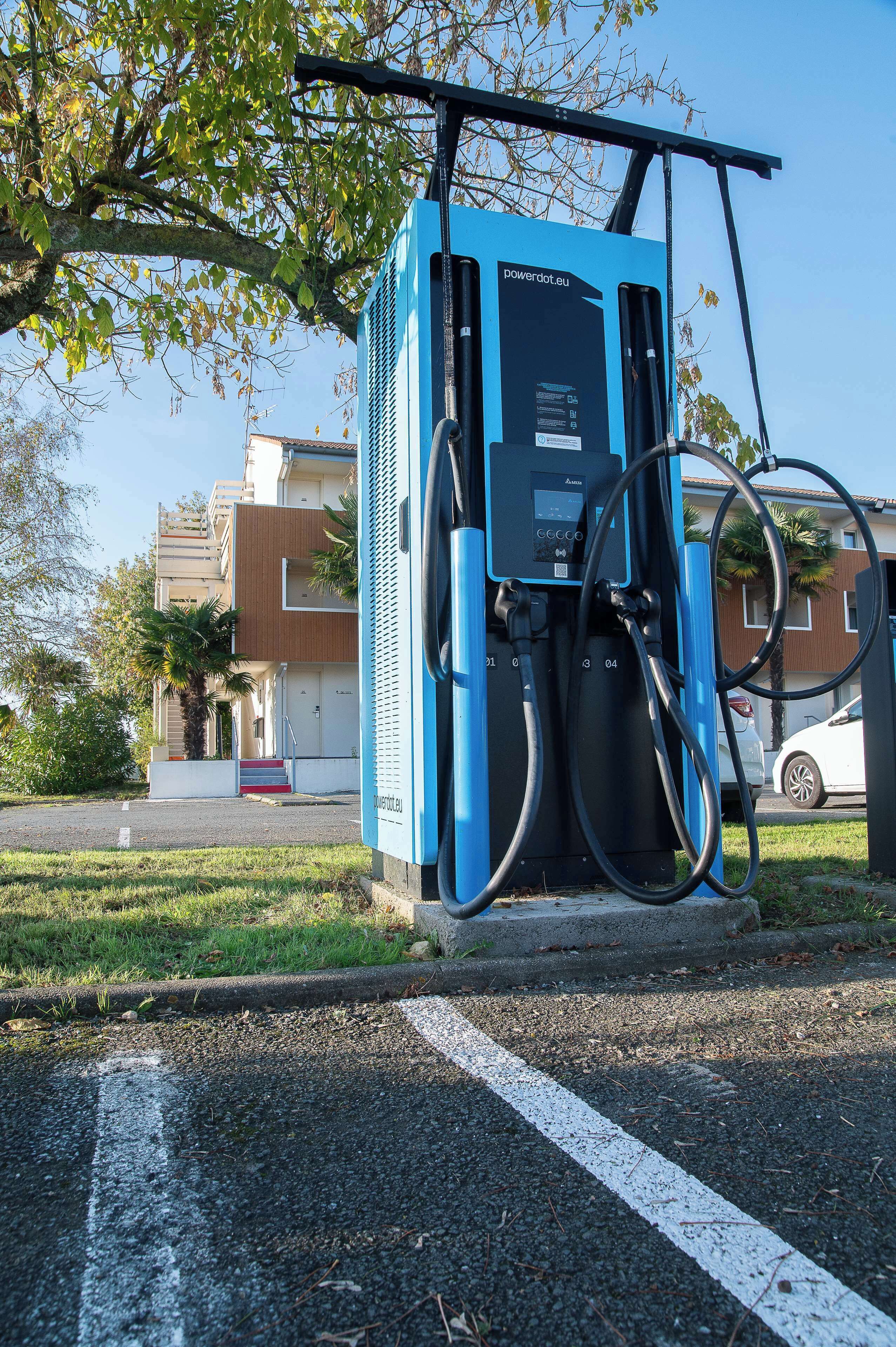Una estación de carga para vehículos eléctricos azules con dos cables, una palmera y un edificio al fondo.