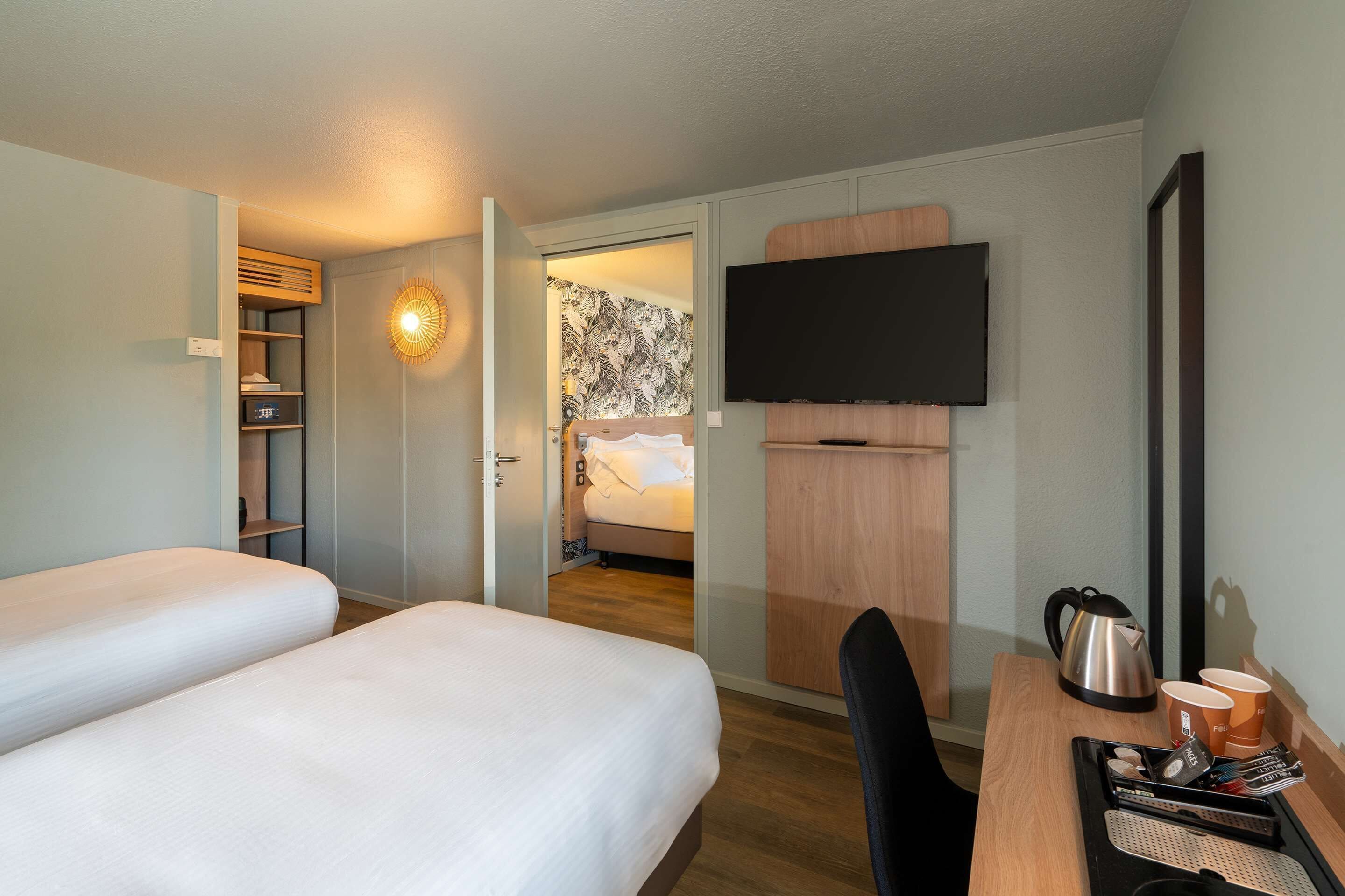 A hotel room with two beds, a desk with a kettle, cups, and a TV mounted on the wall.
