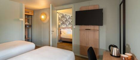 A hotel room with two beds, a desk with a kettle, cups, and a TV mounted on the wall.