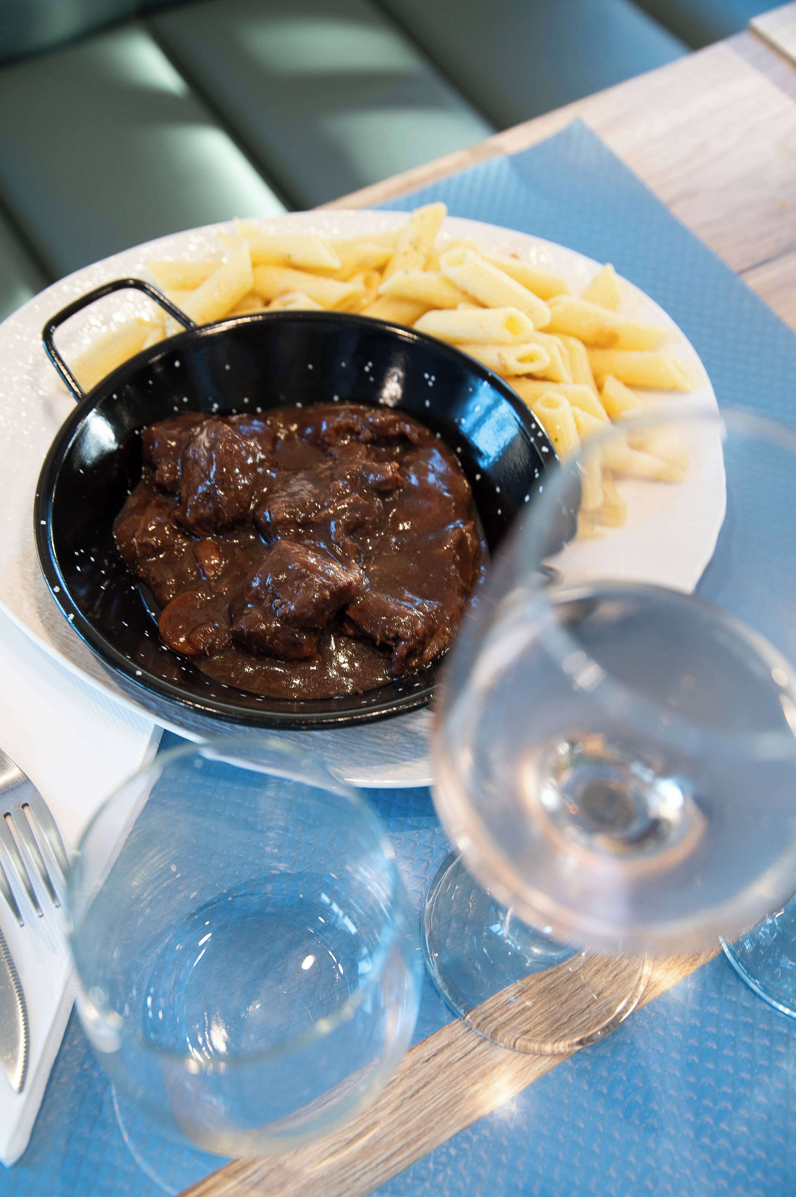 A plate with a bowl of beef stew and pasta, a glass of wine, and a fork on a table with a blue placemat.