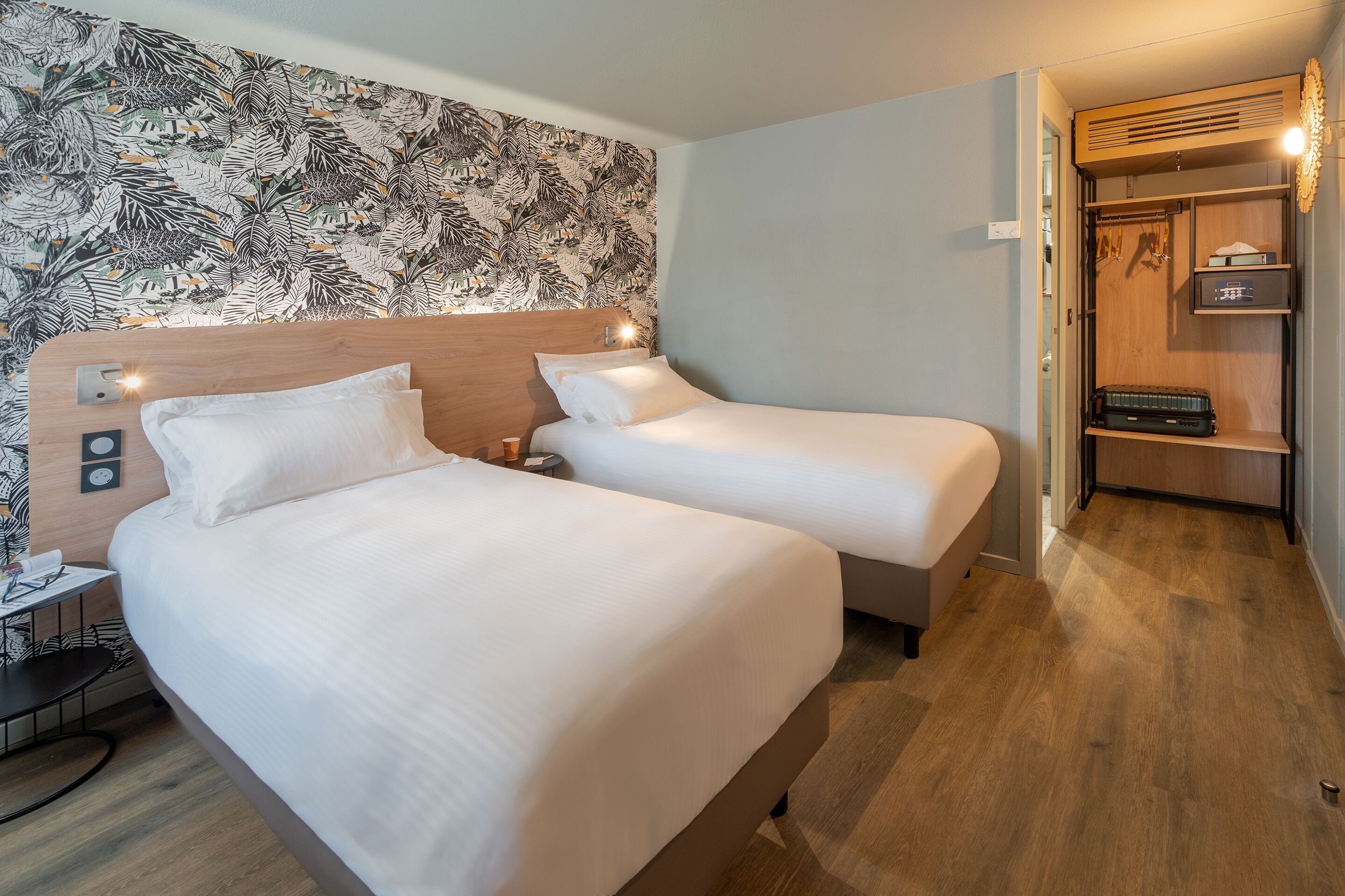 Hotelkamer met twee bedden, een houten hoofdbord, een tafeltje met een stoel, een kledingkast en een behang met patroon.
