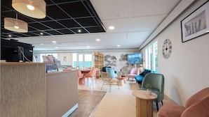 A reception area with a wooden reception desk, a television, and a clock on the wall. There are several seating areas with chairs and tables, and a large window providing natural light. - Sure Hotel by Best Western Rochefort-Sur-Mer (Tonnay-Charente)