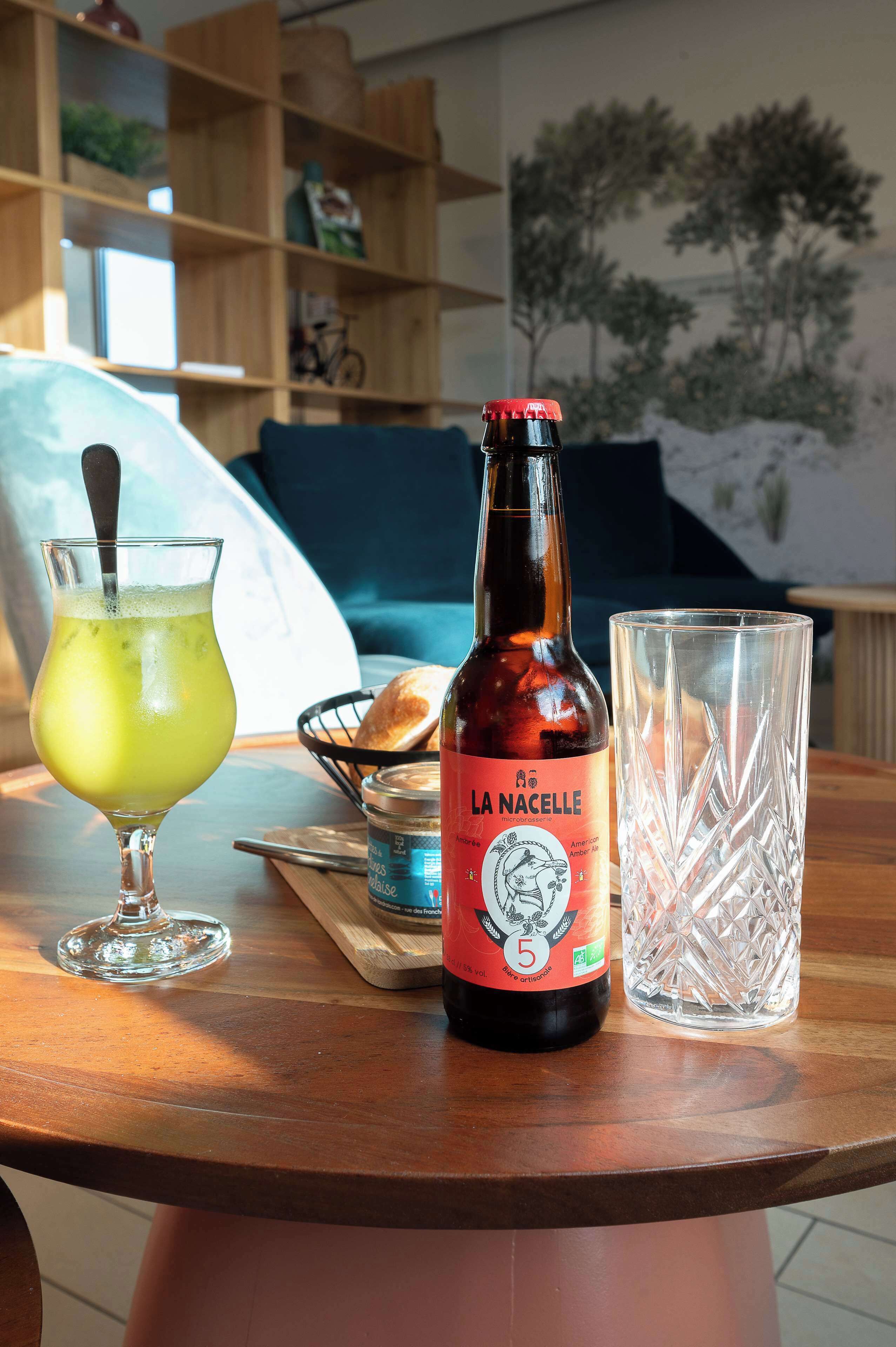 Una botella de cerveza La Nacelle, un vaso de bebida verde y un vaso transparente sobre una mesa de madera.