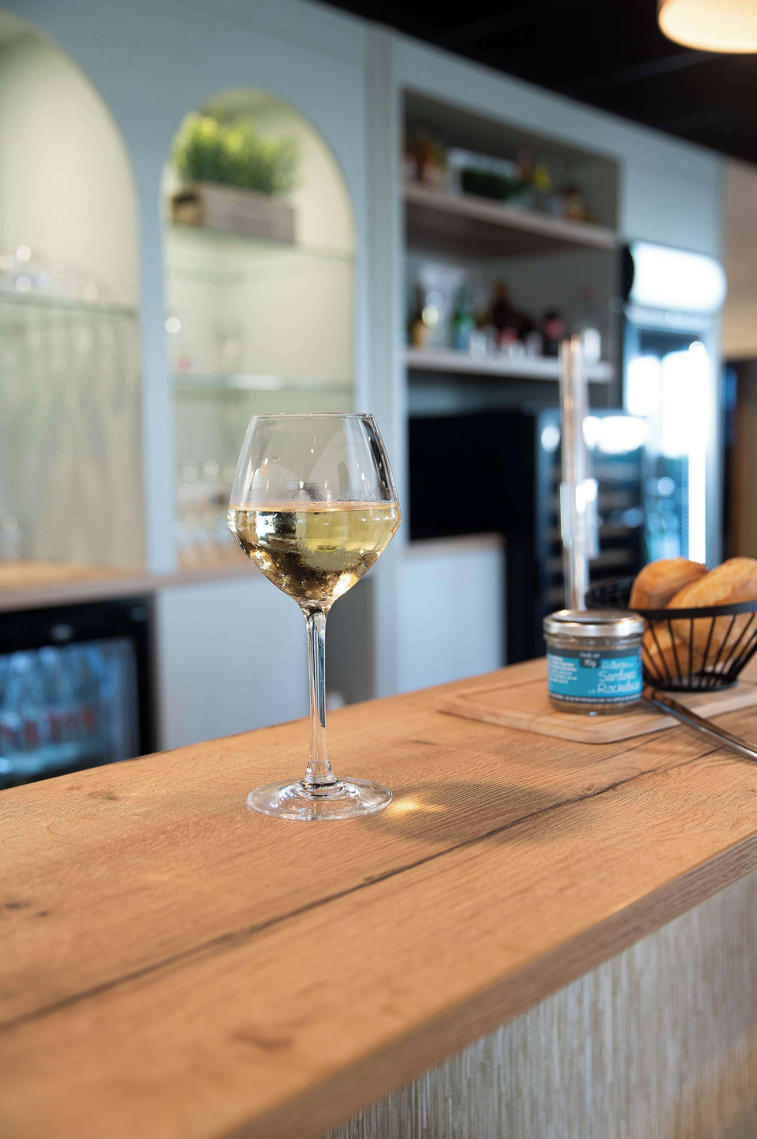 Una copa de vino blanco sobre una barra de madera, con una lata de cerveza y una cesta de pan al fondo.