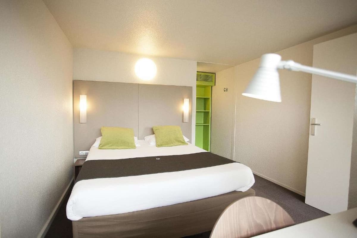 A hotel room with a bed, a bedside lamp, a nightstand, and a green storage unit.