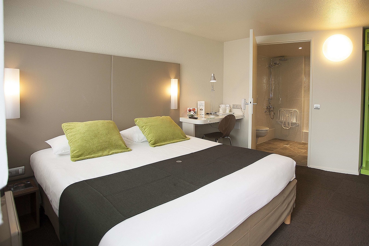 Quarto de hotel com uma cama grande, uma escrivaninha com cadeira, área de chuveiro e uma porta verde.