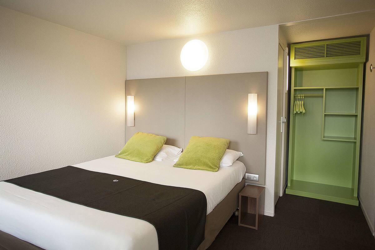 A hotel room with a bed, a nightstand, a green door, and a wall-mounted light fixture.
