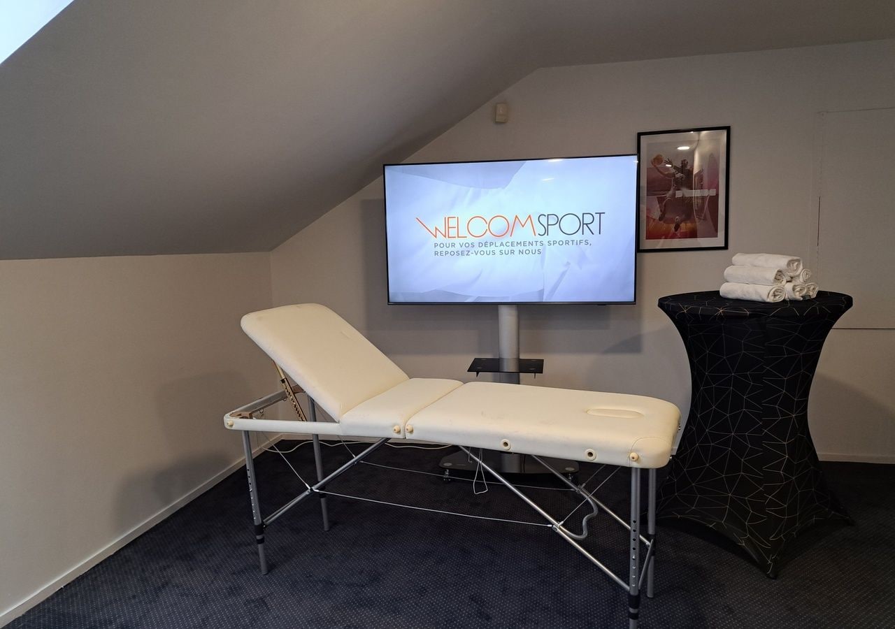 A room with a reclining chair, a television displaying 'WELCOME SPORT' text, a framed picture on the wall, and a small table with towels.