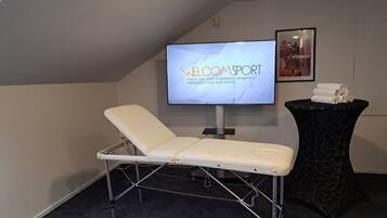 Una habitación con una silla reclinable, un televisor mostrando el texto 'WELCOME SPORT', un cuadro en la pared y una mesita con toallas.