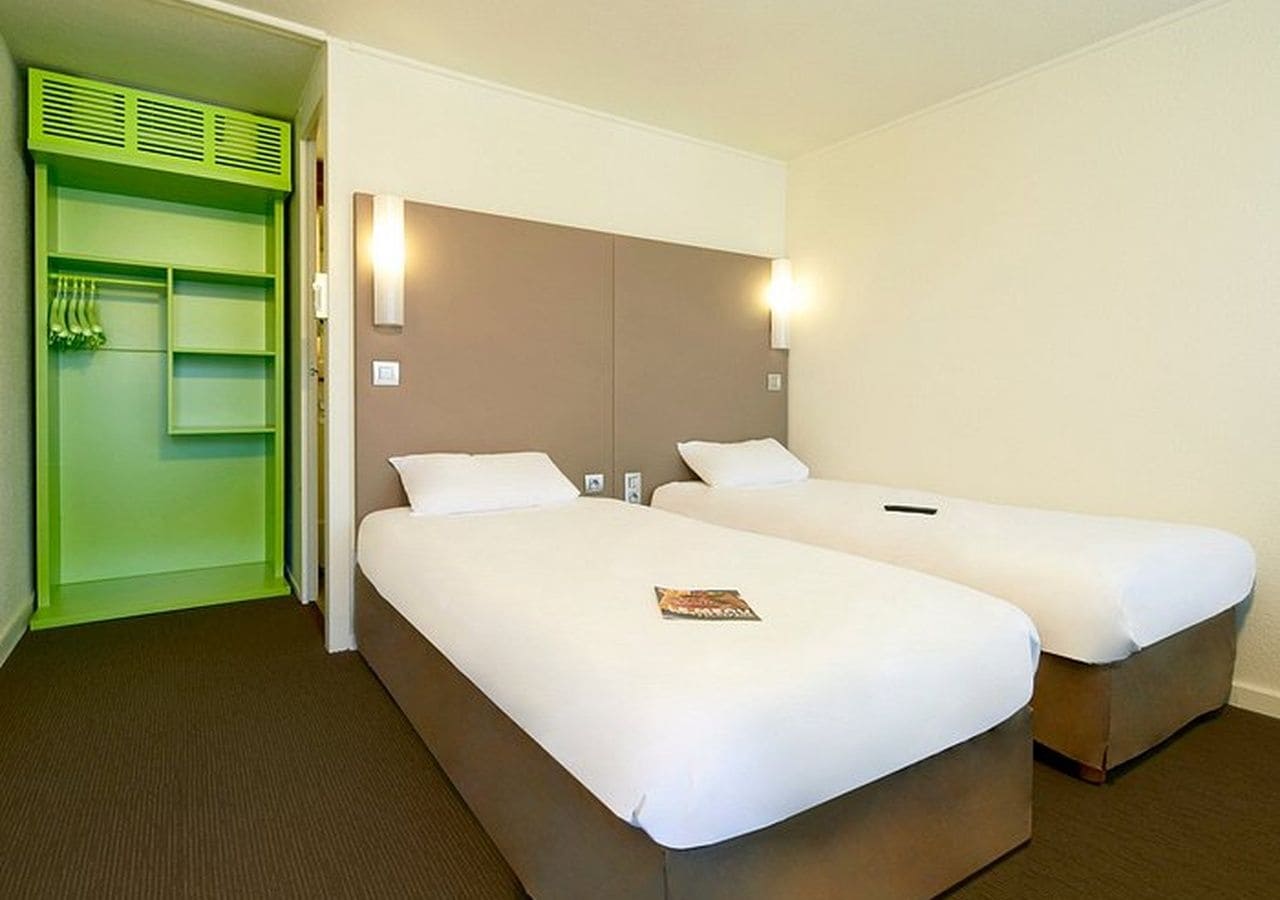 A hotel room with two beds, a green wardrobe, and a light-colored headboard.