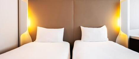 Habitación de hotel con dos camas individuales, cada una con colchón y almohada blancos, y cabecero con iluminación integrada en la pared.