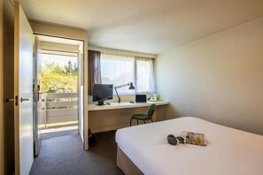 Ein Hotelzimmer mit einem Bett, einem Schreibtisch mit Computer, einem Sessel und einem Fenster mit Vorhängen.