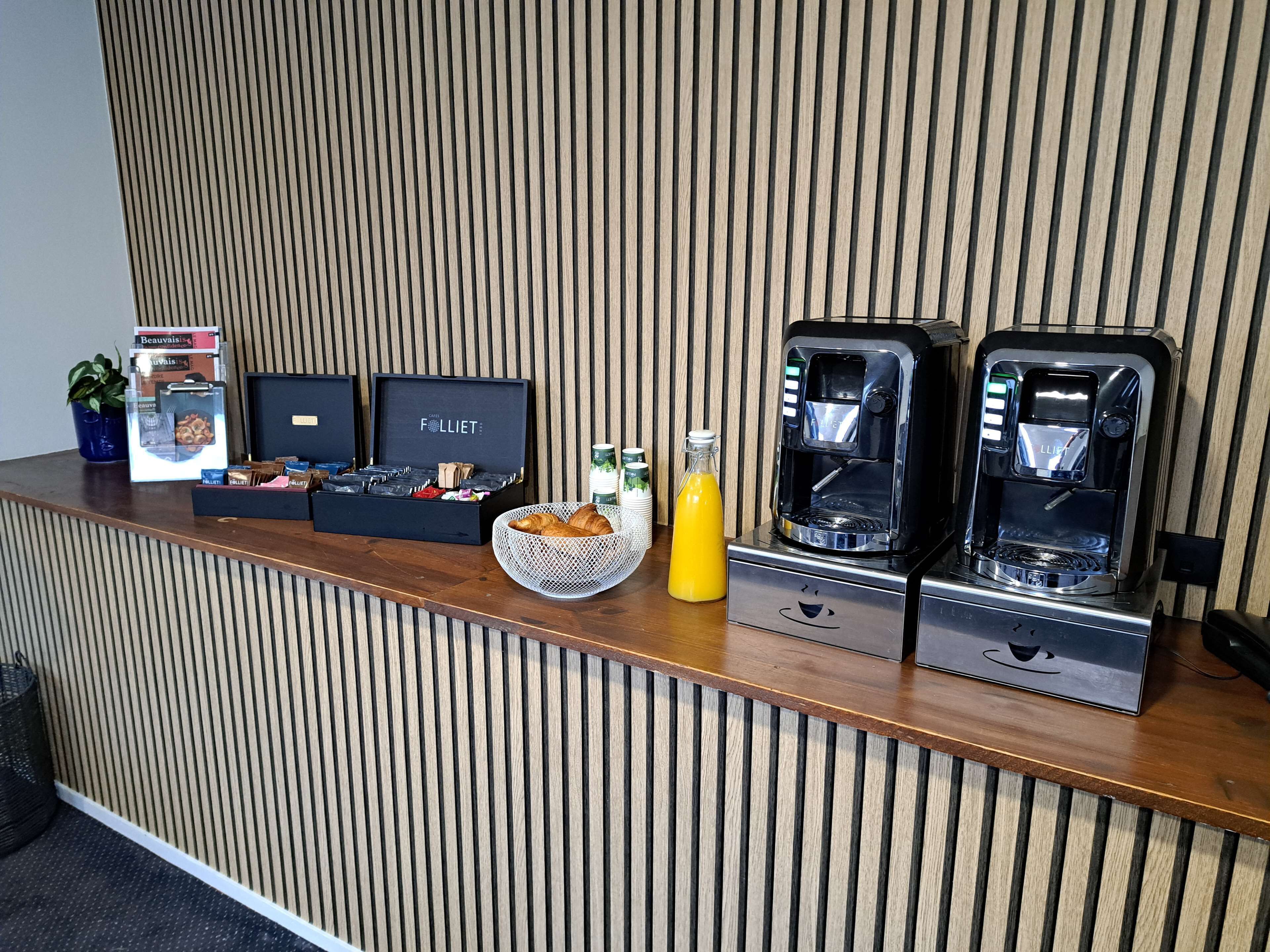 A reception area with a wooden panel wall, a coffee machine, a basket of croissants, a bottle of orange juice, and a box of chocolates.