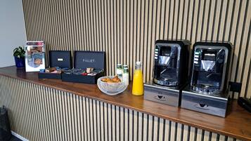Zona de recepción con un panel de madera, una máquina de café, una cesta de cruasanes, una botella de zumo de naranja y una caja de bombones.