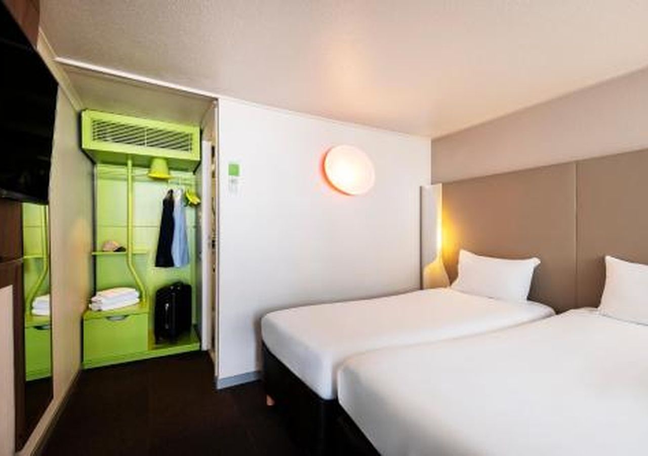A hotel room with two beds, a green wardrobe, and a wall-mounted light fixture.