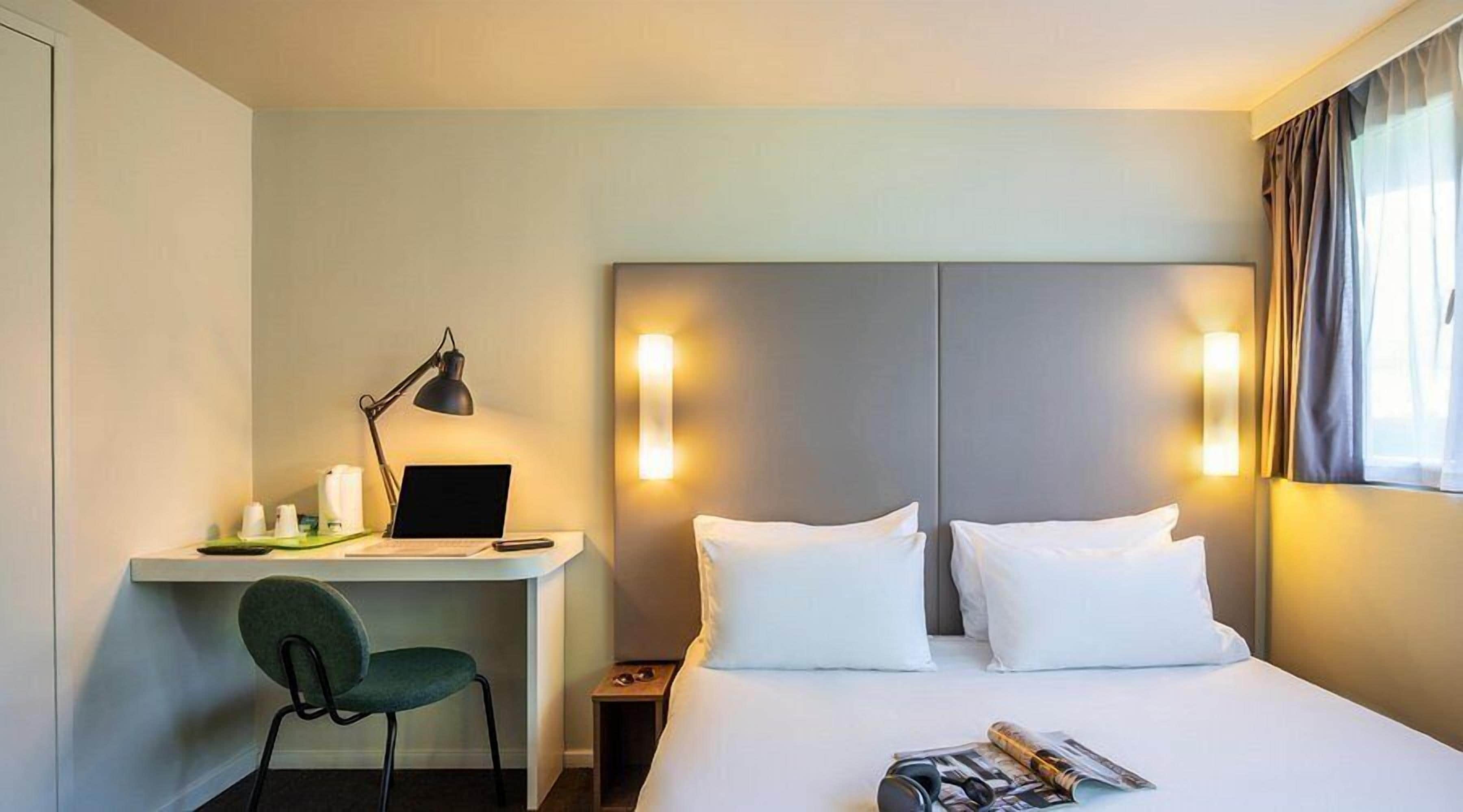 Ein Hotelzimmer mit einem Bett, einem Schreibtisch mit Computer, einem Stuhl und einer Lampe.