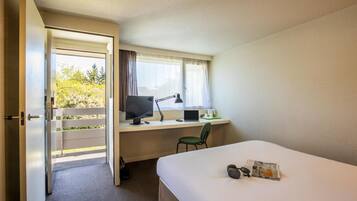 A hotel room with a bed, a desk with a computer and a chair, a window with curtains, and a door leading to an outdoor area.