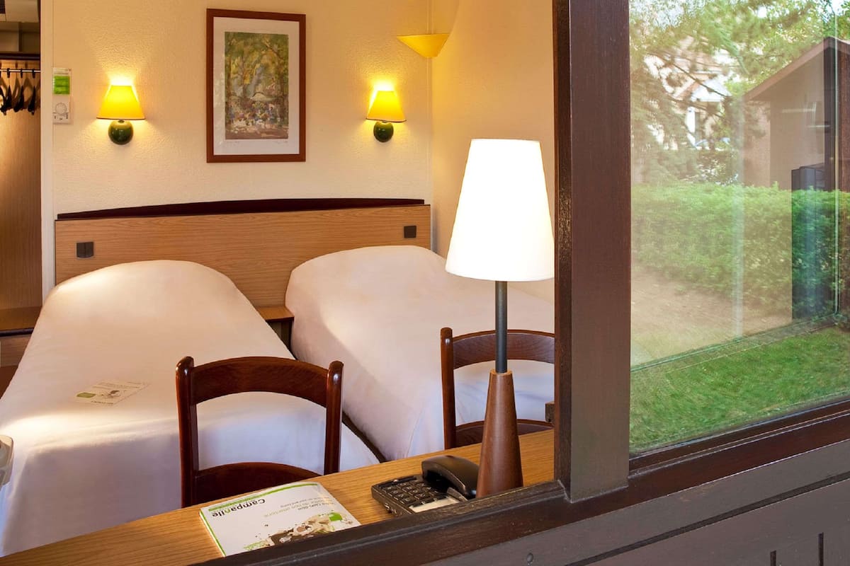 Habitación de hotel con dos camas, un escritorio con lámpara, un cuadro en la pared y una ventana con vistas a zonas verdes.