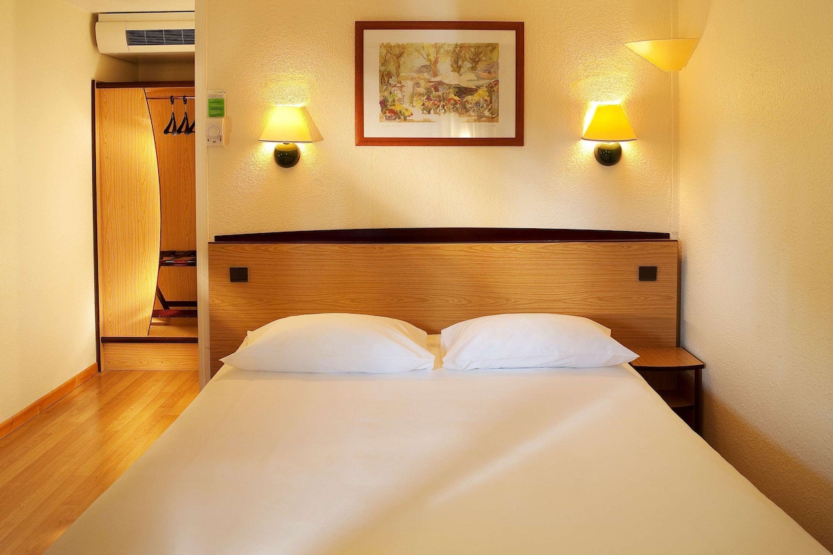 Habitación de hotel con cabecera de madera, dos lámparas de noche, una obra de arte enmarcada en la pared y un armario con ropa colgada.