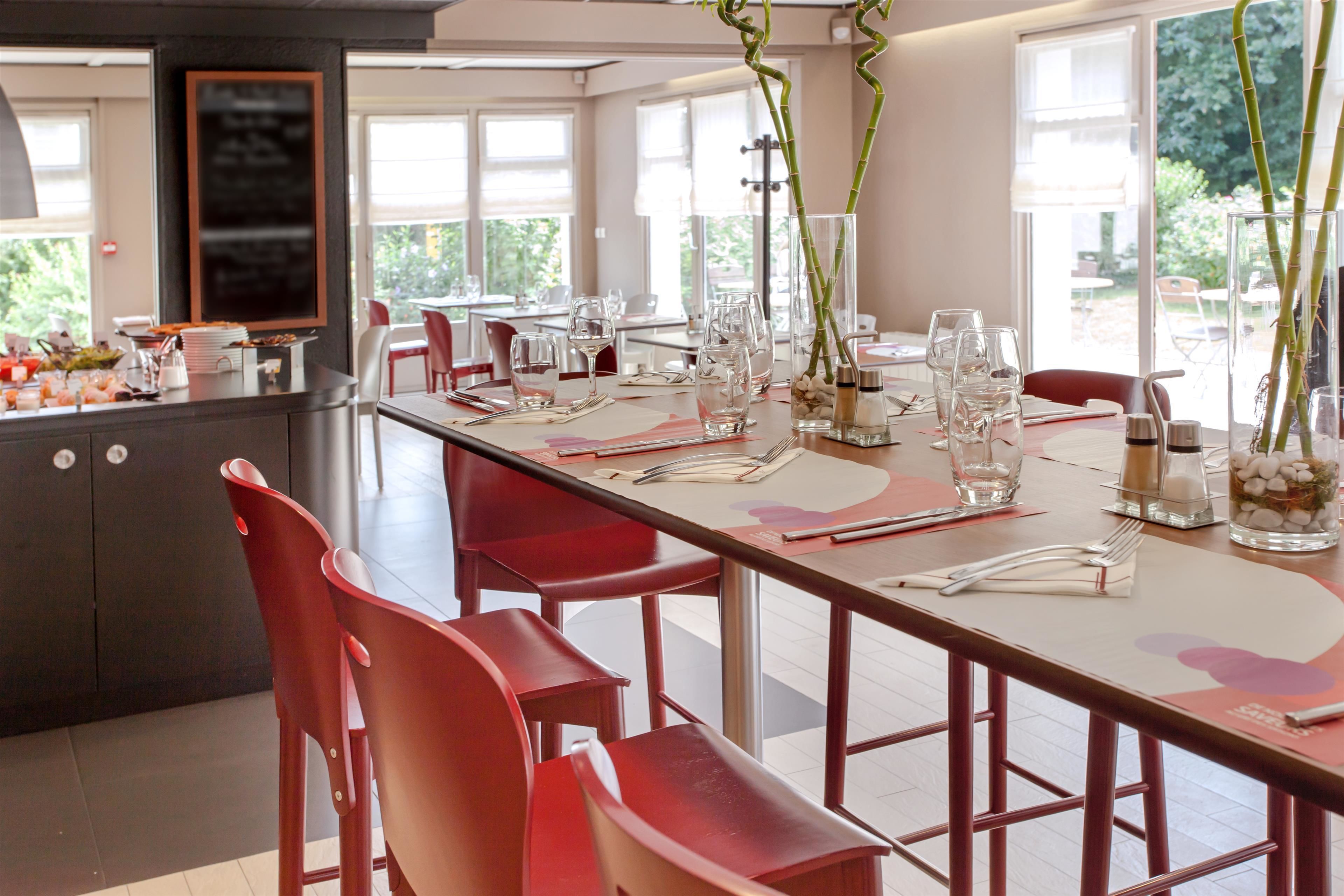 A restaurant with a long dining table set for a meal, red chairs, and a view of the outdoors through large windows.