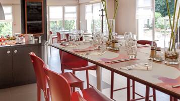 A restaurant with a long dining table set for a meal, red chairs, and a view of the outdoors through large windows.