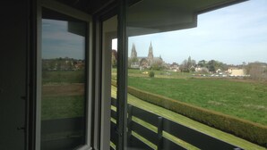 A balcony with a view of a green field and a distant church tower. - Campanile Bayeux (Bayeux)