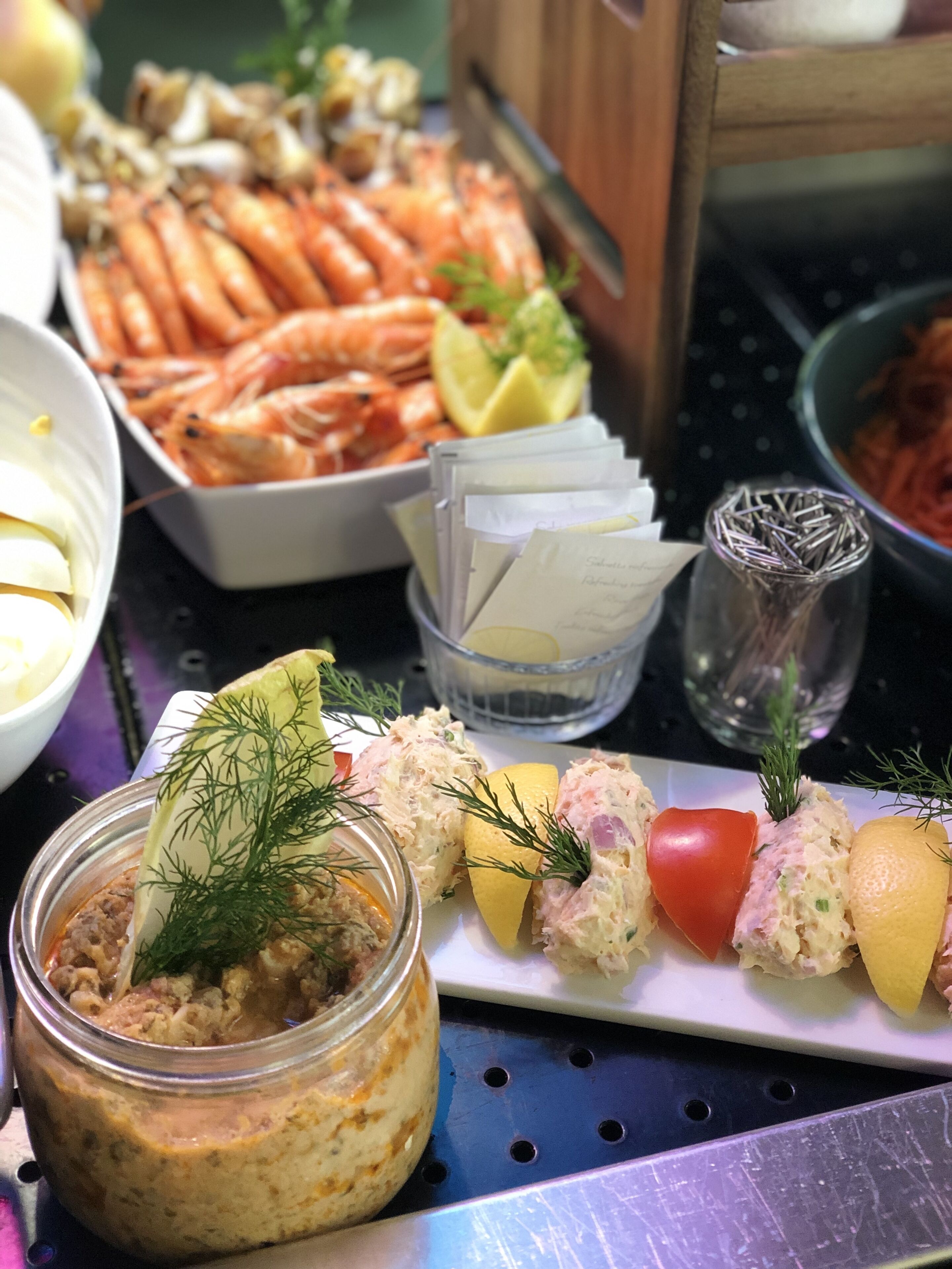 Une table de buffet proposant divers plats, notamment des crevettes, un bocal de salade et une assiette décorée de tomates et de citron.