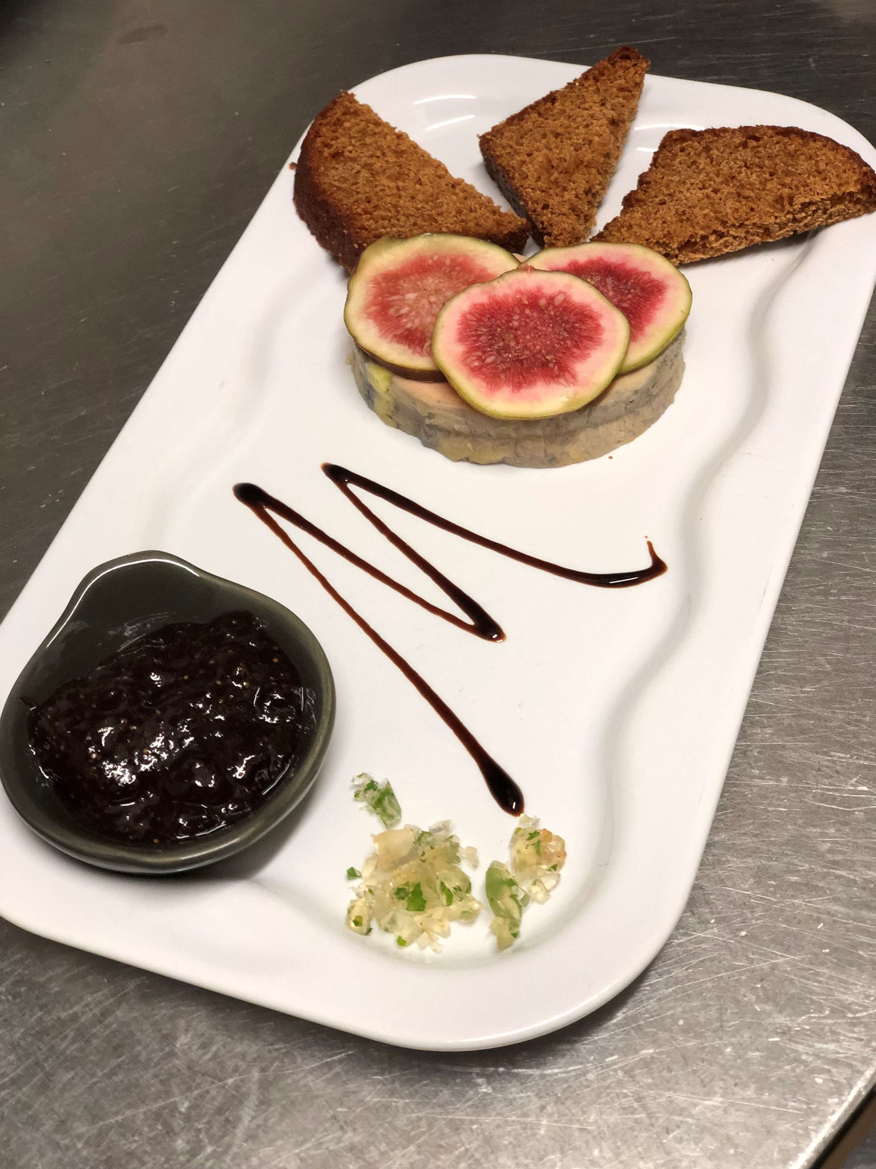 Une assiette rectangulaire blanche sur laquelle est posée une part de gâteau, garnie de tranches de figues, un petit bol de sauce foncée et quelques oignons verts hachés.
