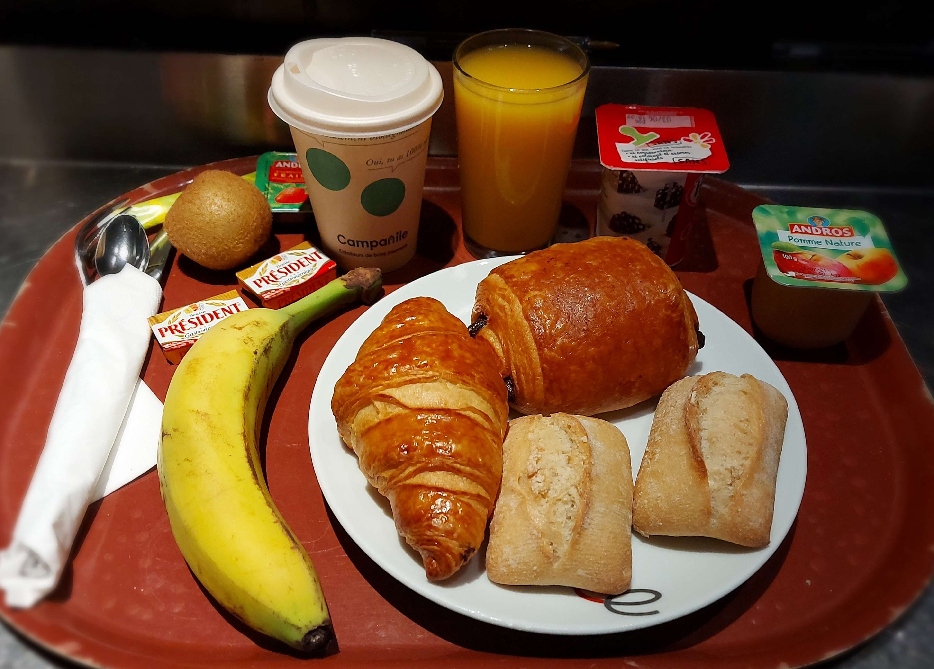 A breakfast tray with croissants, a banana, juice, yogurt, and a coffee cup.