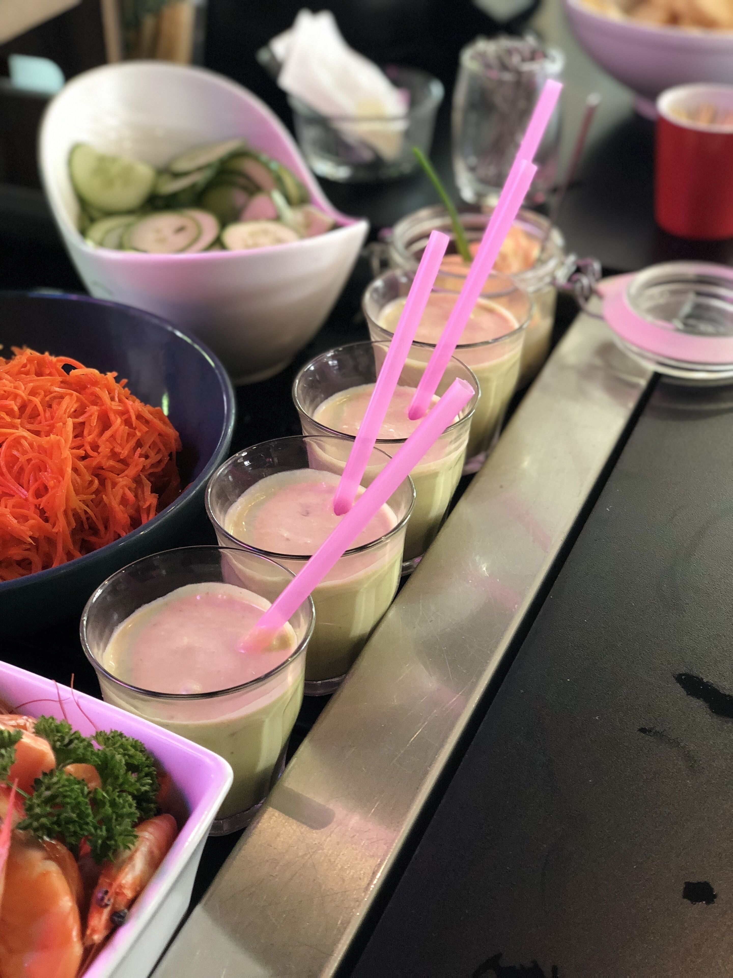 Une table de buffet proposant divers plats, notamment un saladier de concombres en tranches, un plat de carottes râpées et une assiette de crevettes garnies de persil.
